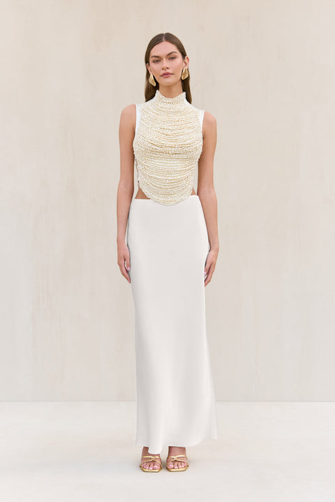 Woman wearing a cropped white tank top that features rows of pearl-beaded strands draped across the bodice, paired with a white maxi skirt. 