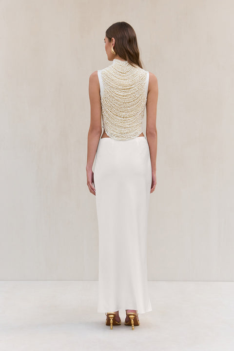 Back-facing woman wearing a cropped white tank top that features rows of pearl-beaded strands draped across the bodice, paired with a white maxi skirt. 