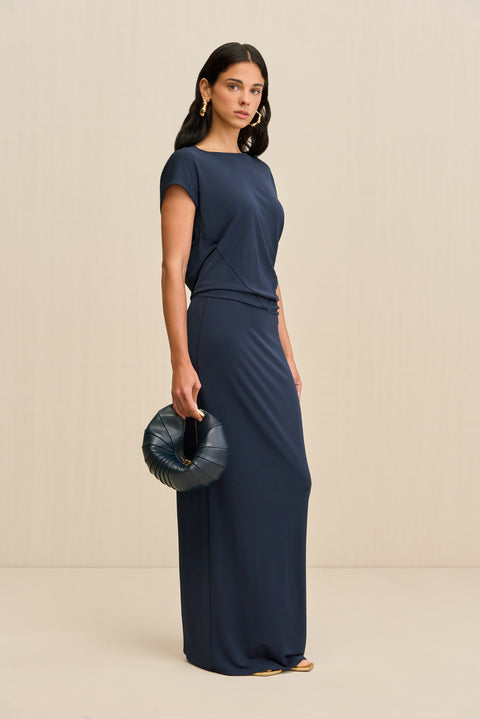 Side-facing woman wearing a slinky navy maxi dress that features an asymmetrical cap sleeve and draping throughout the bodice, paired with a navy handbag and gold sandals. 