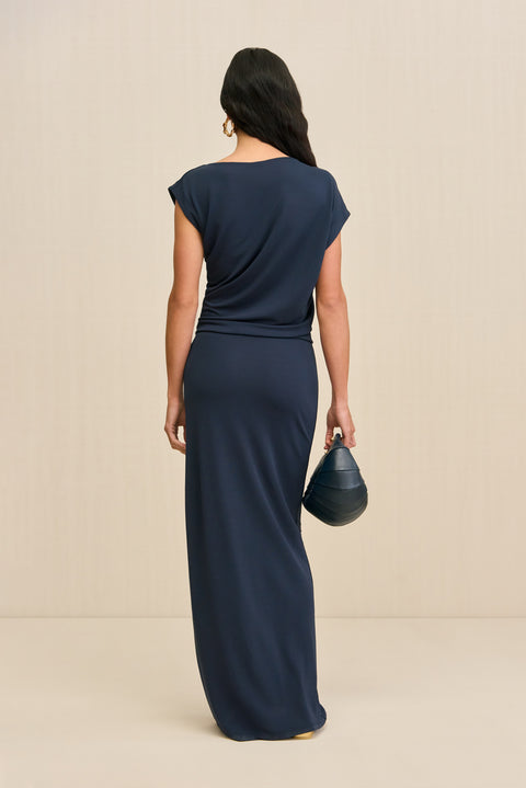 Back-facing woman wearing a slinky navy maxi dress that features an asymmetrical cap sleeve and draping throughout the bodice, paired with a navy handbag and gold sandals. 