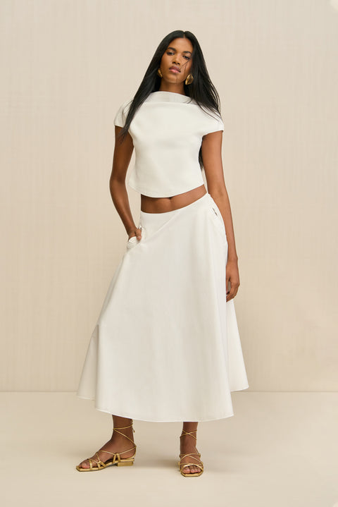 Woman posing and wearing a white poplin, A-line midi skirt paired with a white top and gold sandals. 