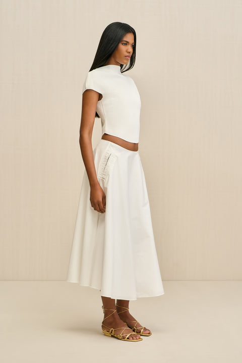 Side-facing woman wearing a white poplin, A-line midi skirt paired with a white top and gold sandals. 