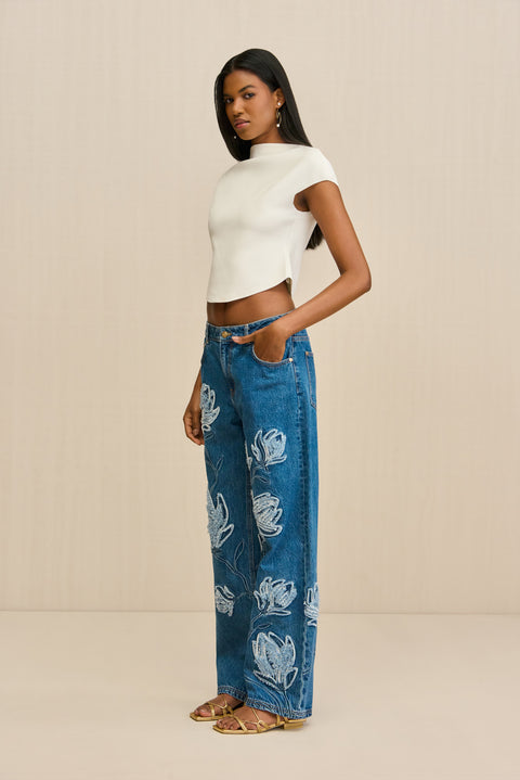 Side-facing woman wearing a pair of blue wash, wide leg jeans that features an embroidered floral design in lighter wash, paired with a white boatneck cropped t-shirt. 