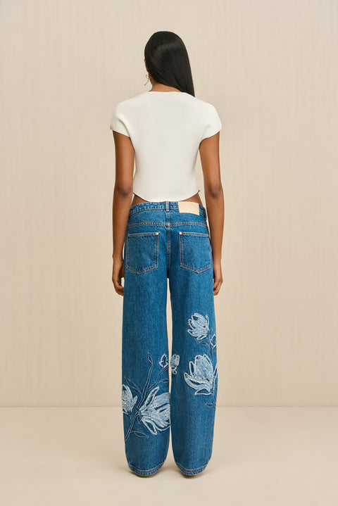 Back-facing Woman wearing a pair of blue wash, wide leg jeans that features an embroidered floral design in lighter wash, paired with a white boatneck cropped t-shirt. 