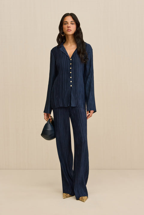 Woman wearing navy pleated pants, paired with a matching button down shirt, gold shoes, and a navy handbag. 