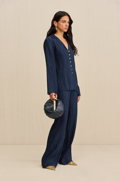 Side-facing woman wearing navy pleated pants, paired with a matching button down shirt, gold shoes, and a navy handbag. 