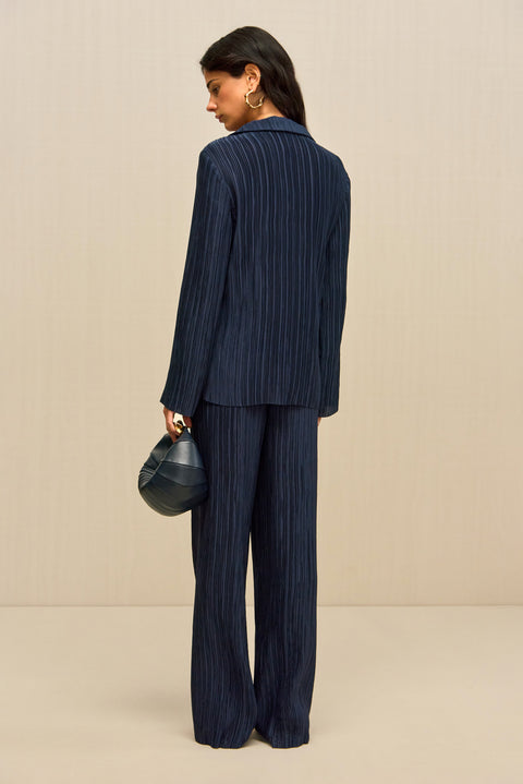 Back-facing woman wearing navy pleated pants, paired with a matching button down shirt, gold shoes, and a navy handbag. 