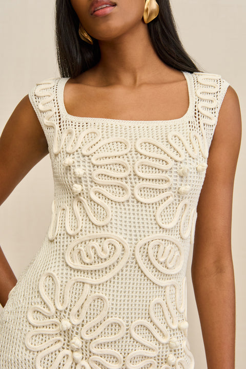 Close up of a woman wearing a sleeveless, white netted mini dress that features a corded floral overlay and square neck with a slip underneath for coverage. 