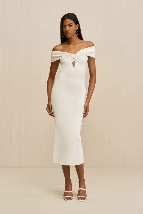 Woman wearing a white knit midi dress that features a twist and keyhole detail at the bust with both straps worn off the shoulders, paired with white sandals. 