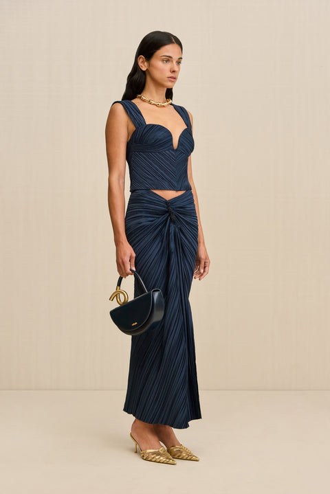 Side-facing woman wearing a navy blue midi skirt that features an asymmetrical waistline with knotted ruching at the hip, paired with a matching top and gold heels. 