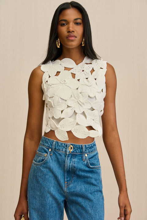 Close up of a woman wearing a white crochet tank top that is crafted from floral appliques sewn together, paired with blue jeans. 