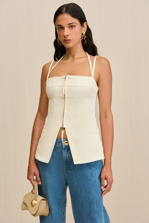 Close up of a woman wearing a white tank top that features a double strap detail, and front center zipper, and a flared cutout at the waist, paired with jeans. 
