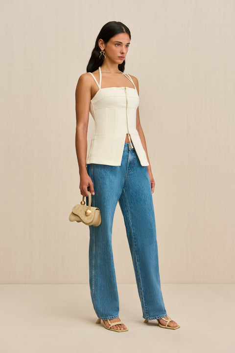 Side-facing woman wearing a white tank top that features a double strap detail, and front center zipper, and a flared cutout at the waist, paired with jeans and sandals. 