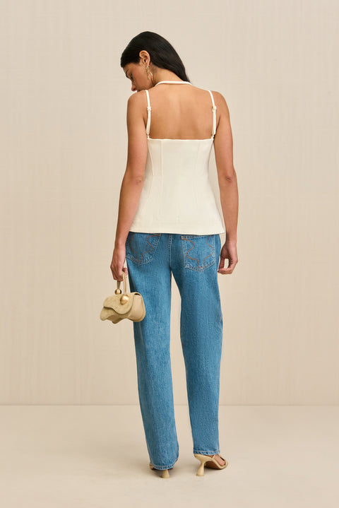 Back-facing woman wearing a white tank top that features a double strap detail, and front center zipper, and a flared cutout at the waist, paired with jeans and sandals. 