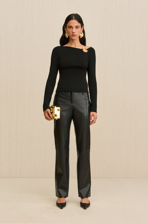 Woman wearing a black long sleeve shirt with a gold ring detail at the shoulder with black leather pants. 