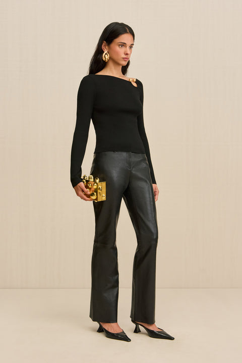 Side-facing woman wearing a black long sleeve shirt with a gold ring detail at the shoulder with black leather pants. 