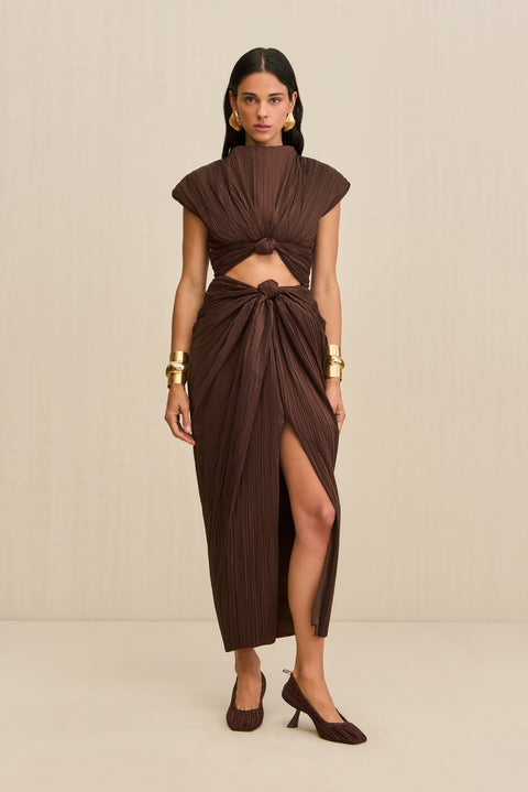 Woman wearing a brown structured midi dress crafted from plisse fabric that features a mock neck, front slit, and a knotted cutout detail below the bust, paired with brown heels. 
