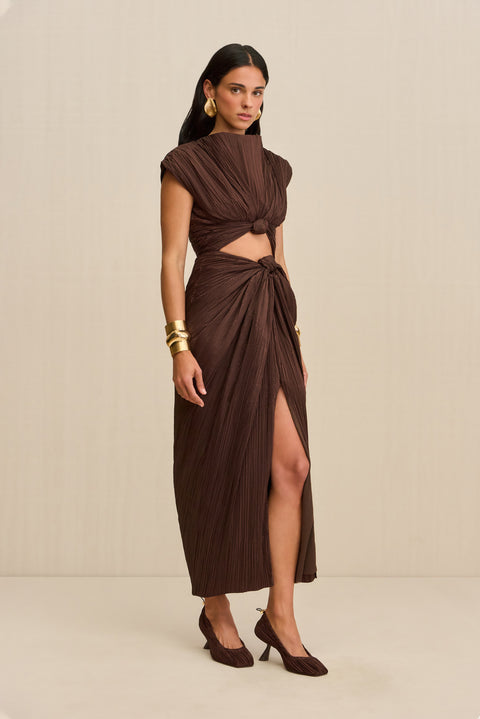 Side view of a woman wearing a brown structured midi dress crafted from plisse fabric that features a mock neck, front slit, and a knotted cutout detail below the bust, paired with brown heels. 