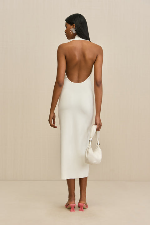 Woman wearing a white midi dress that features a  halter neck and open back, paired with a white handbag and pink heels. 