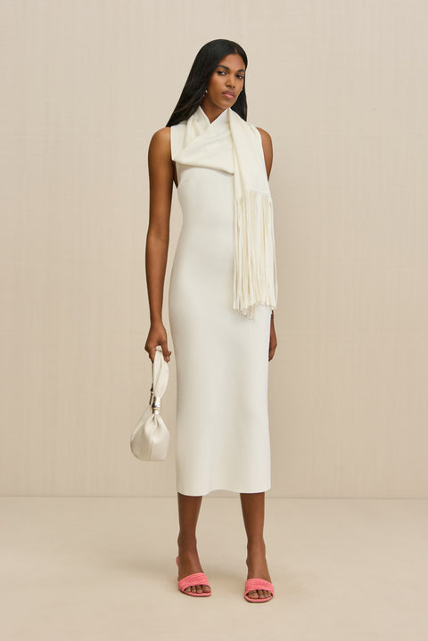 Woman wearing a sleeveless, white knit midi dress that features a removable scarf, paired with pink heels and a white handbag. 