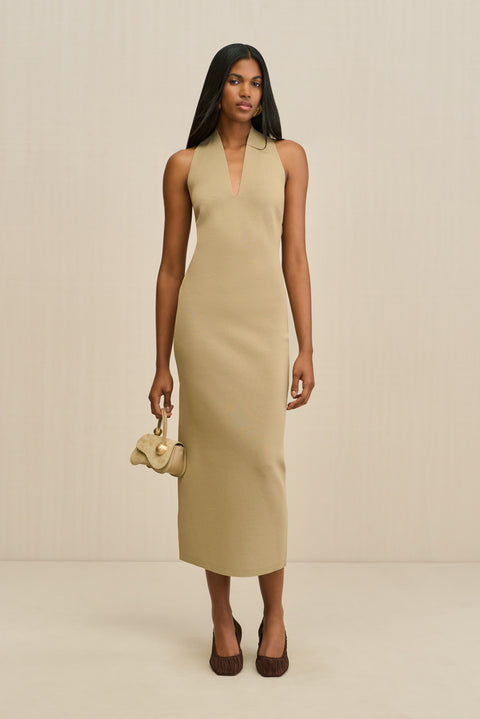 Woman wearing a tan, knit midi dress that features a V neck and a detachable fringe scarf, paired with a gold handbag and brown heels. 