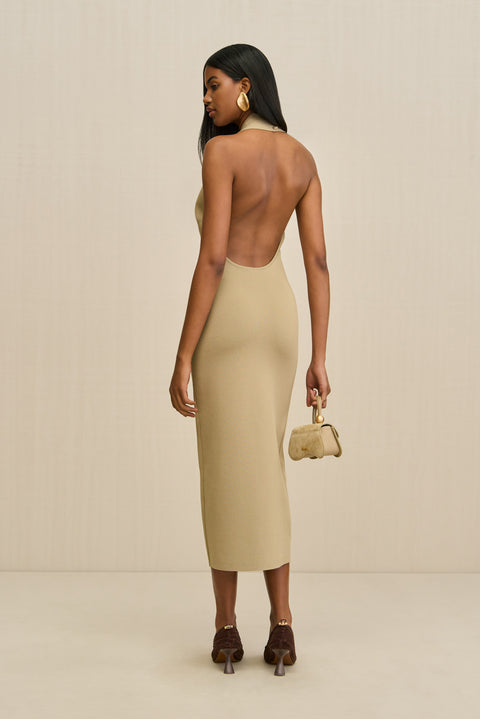 Back-facing woman wearing a tan knit midi dress that features an open back and a halter neck, paired with a gold handbag and brown heels. 