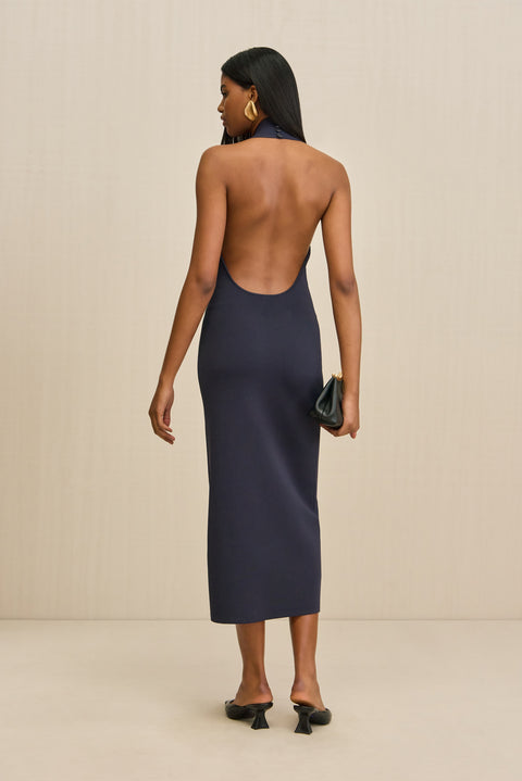 Back-facing woman wearing a navy sleeveless midi dress that features a halter neckline and open back, paired with black sandals. 