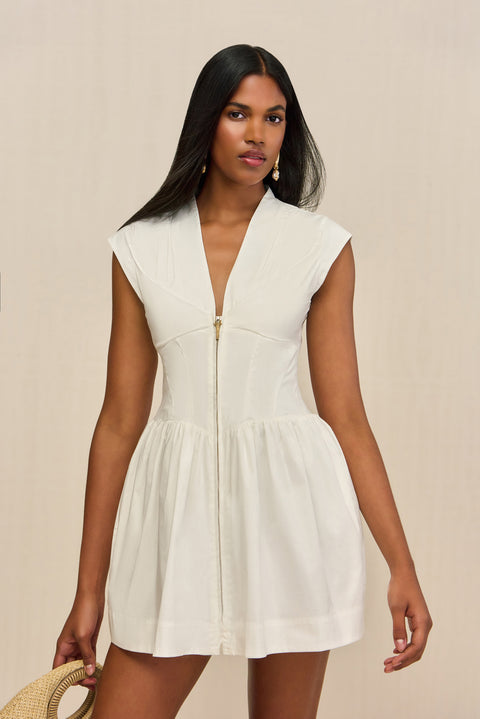 Close up of a woman wearing a white mini dress that features a Deep V, front center zipper, and fitted waist, paired with a tan handbag. 