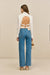 Back view of a woman wearing a white button down that features an open back secured by two adjustable buckles, paired with blue jeans and tan sandals. 