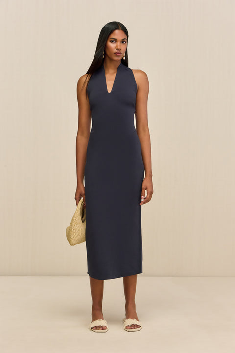 Woman wearing a navy sleeveless midi dress that features a halter V-neck and form-fitting silhouette paired with cream sandals. 