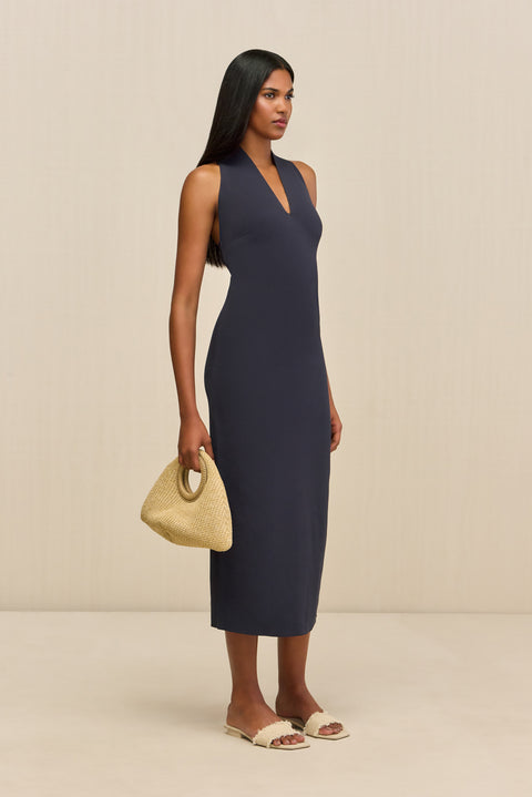 Side-facing woman wearing a navy sleeveless midi dress that features a halter V-neck and form-fitting silhouette paired with cream sandals. 