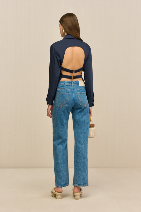 Back-facing woman wearing a navy collared shirt that features an open back secured by two adjustable buckle straps , paired with blue jeans and tan sandals. 