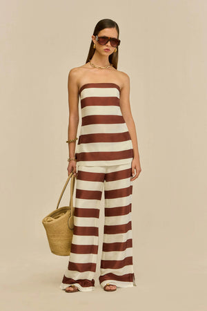 Woman wearing a strapless brown and white striped top and wide leg pant holding tote bag against a beige background