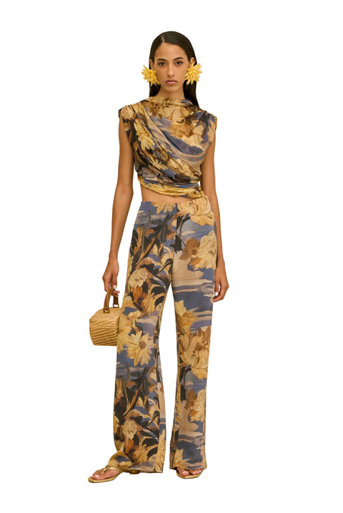 Woman wearing a floral jumpsuit on a beige background holding a woven bag in one hand.