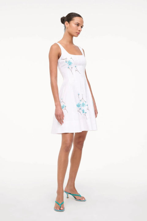 Side-facing woman wearing a sleeveless white mini dress that features a square neck, corset-style paneling, an A-line skirt, and blue beaded dandelion details throughout. 