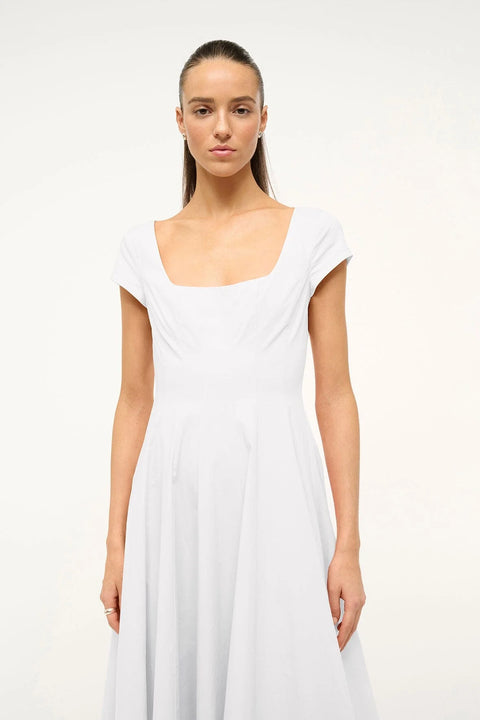 Close up of a woman wearing a white midi dress that features cap sleeves, a square neck, corset styles paneling, and an A-line skirt.