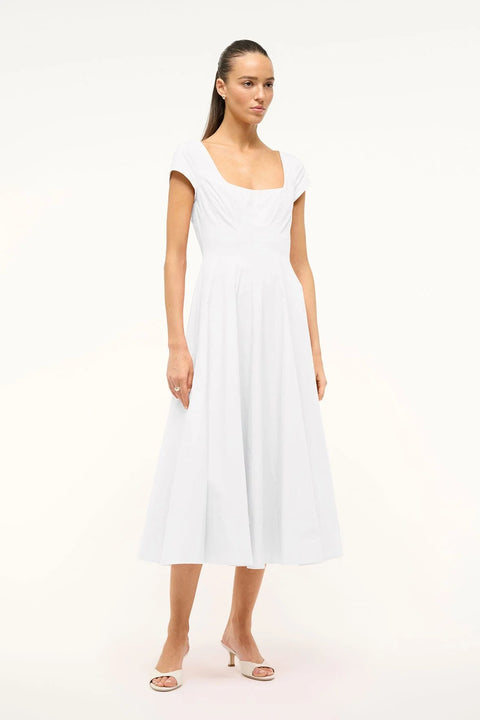 Side view of a woman wearing a white midi dress that features cap sleeves, a square neck, corset styles paneling, and an A-line skirt, paired with white heels. 