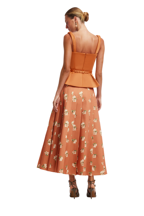 A woman shown from the back wearing a sleeveless orange-brown set with a structured peplum waist and a flared skirt featuring a small floral print, paired with brown lace-up heels against a plain white background.