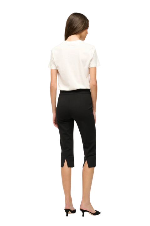 A woman shown from the back wearing a white short-sleeve top, black cropped capri pants with side slits, and black pointed mule heels against a plain white background.