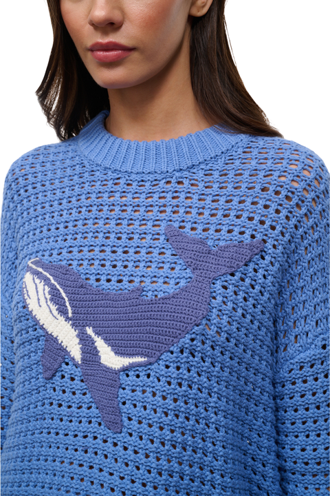 A close-up of a woman wearing a blue open-knit sweater with a large purple and white whale motif across the chest.