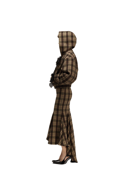 A model shown in profile wearing a brown and black plaid hooded jacket and matching long skirt, paired with black high heels, against a solid white background.