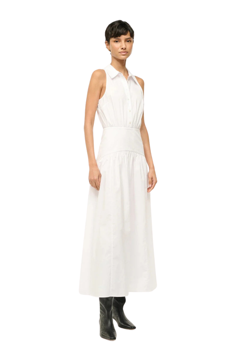 A model wearing a sleeveless white midi dress with a collared neckline and gathered waist, paired with black ankle boots, set against a plain white background.
