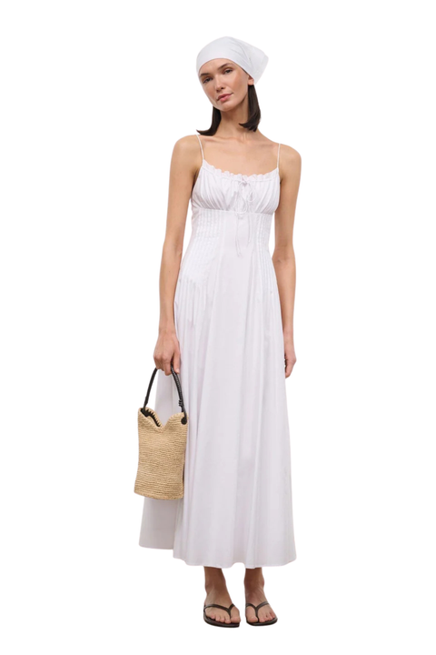 A woman modeling a white sleeveless maxi dress with a smocked bodice, paired with a white headscarf, straw bucket bag, and brown sandals, set against a plain white background.