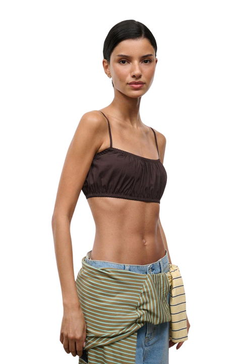 A model wearing a dark brown ruched spaghetti-strap crop top, styled with a green and white striped sarong tied around the waist over light-wash blue jeans, against a solid white background.