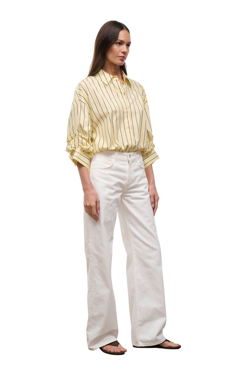 A woman modeling a yellow and white vertically striped button-down shirt with rolled-up sleeves, paired with off-white wide-leg trousers and dark sandals, set against a plain white background.