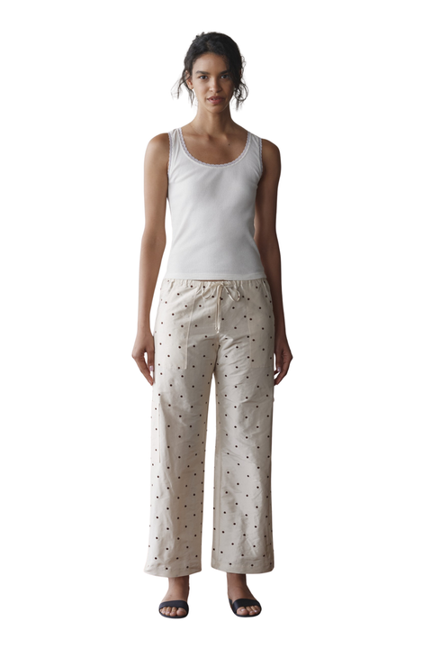 A woman modeling a two-piece loungewear set consisting of a white tank top and cream-colored wide-leg pants with a small black pattern, shot against a solid white background.