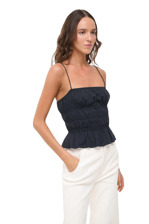 A woman modeling a black ruched spaghetti-strap top paired with white pants, photographed against a solid white background.