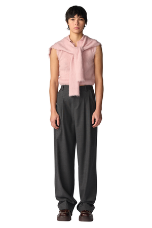 A model stands against a white background wearing a light pink sleeveless fuzzy top with a draped scarf detail, paired with dark gray pleated trousers and brown shoes.