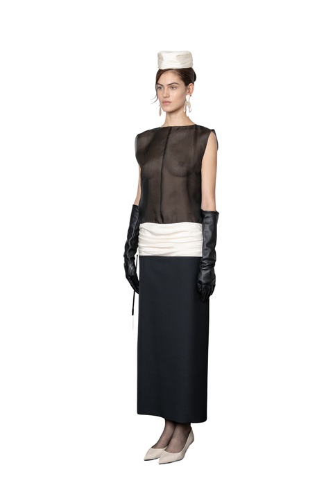 A model is shown wearing a vintage-inspired black and white outfit consisting of a sheer black top, a white waist sash, a long black skirt, black leather opera gloves, and a small white hat, set against a plain white background.
