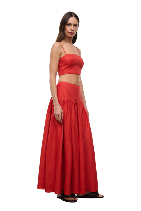 A woman modeling a bright red two-piece outfit consisting of a sleeveless crop top with thin straps and a matching high-waisted, flowing maxi skirt, set against a plain white background.